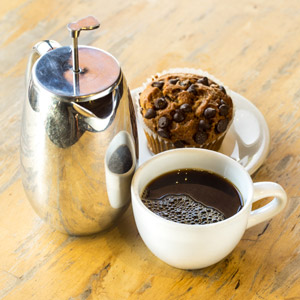 silver french press next to cup of coffee and muffin